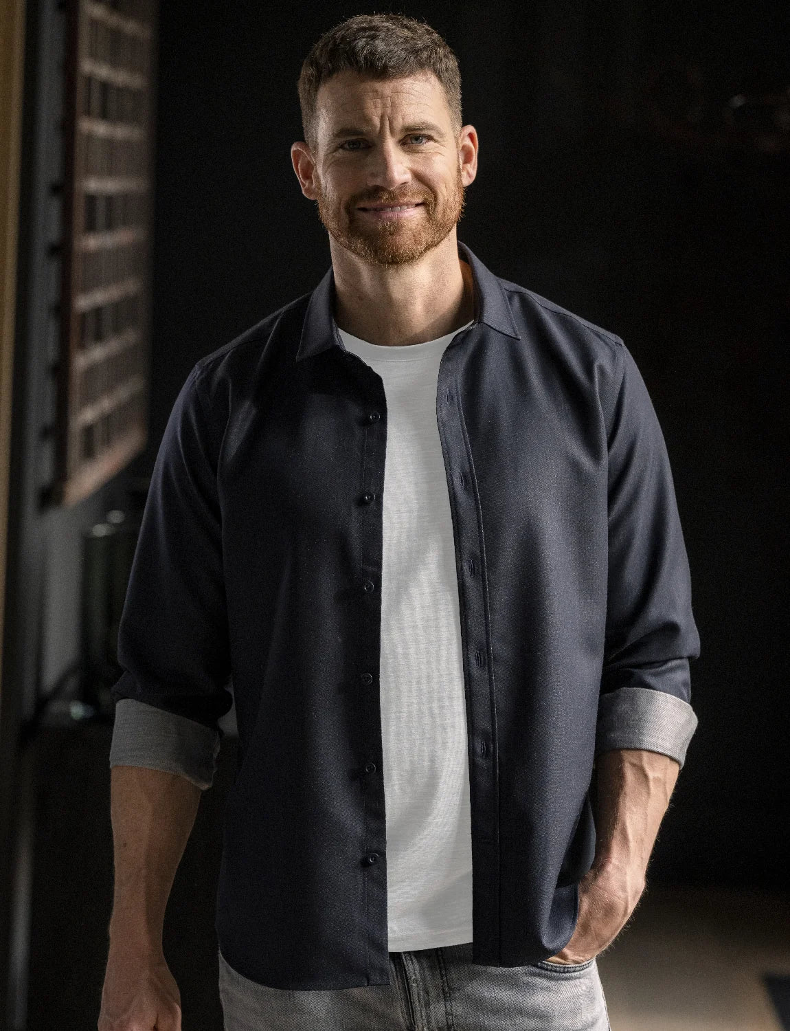 A man wearing a dark navy blue button-down shirt over a white t-shirt and light gray jeans, standing in a dimly lit indoor setting.