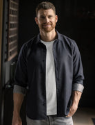 A man wearing a dark navy blue button-down shirt over a white t-shirt and light gray jeans, standing in a dimly lit indoor setting.