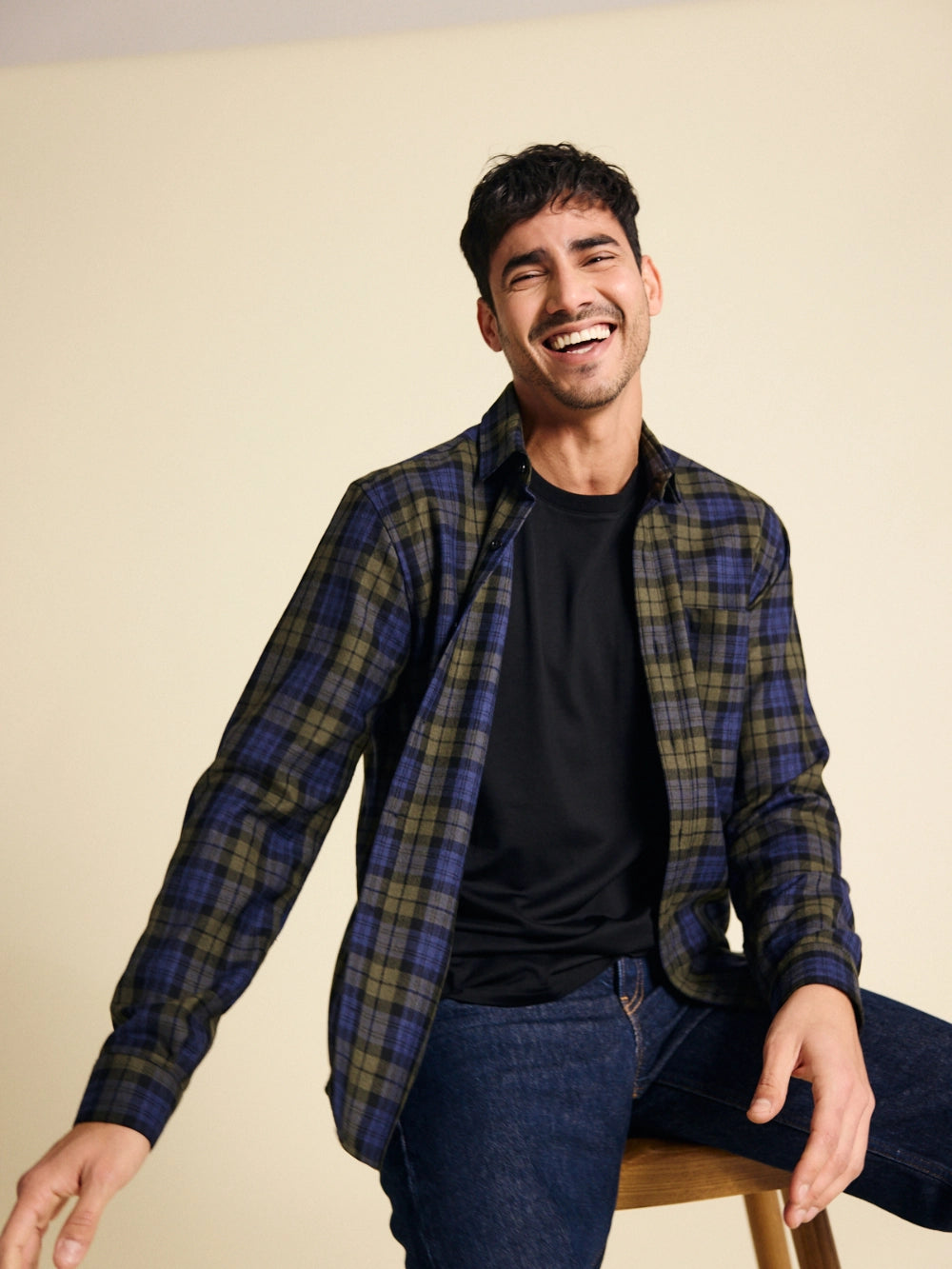 Man wearing a plaid merino flannel shirt over a black t-shirt, sitting on a stool against a beige background