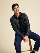 Man wearing a merino wool plaid shirt and jeans sitting on a stool against a beige background