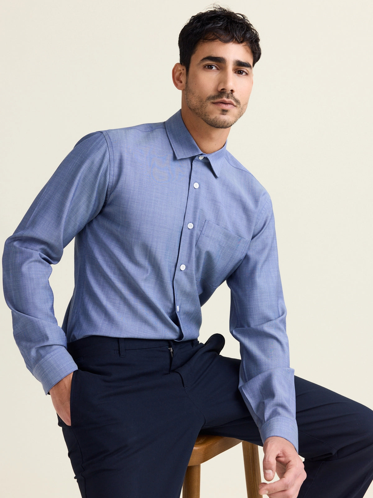 Man sits on a high chair and wears a merino wool shirt in light blue denim color with chest pocket and white buttons tucked in on a dark navy pants