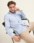 Man sitting on a chair wearing a light blue striped merino wool shirt with chest pocket from Wolk and beige pants.