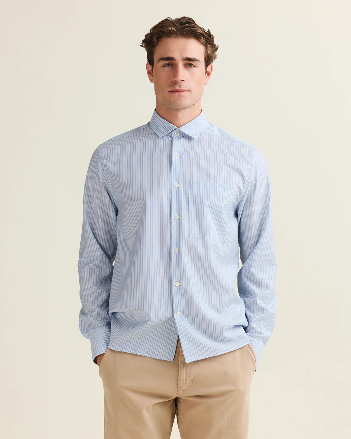 Man wearing a light blue striped merino shirt with chest pocket in relaxed boxy fit and beige pants on a plain background from the brand Wolk