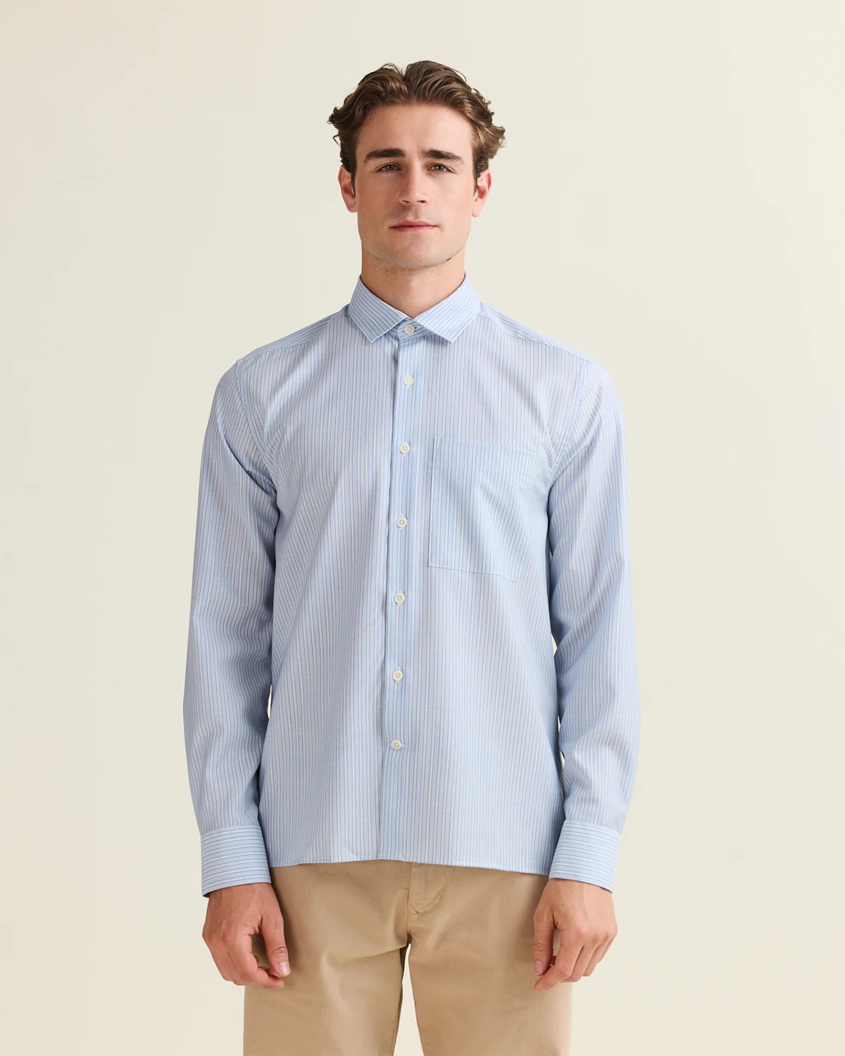 Front view of Man wearing a light blue striped merino shirt with chest pocket, straight hem in boxy fit and beige pants on a plain background