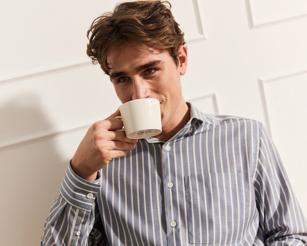 Man drinking from a white mug against a light background wearing a navy stripe merino wool shirts with long sleeves and a chest pocket
