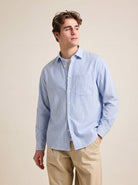 Man wearing a blue striped merino shirt with long sleeves and chest pocket on a beige pants against a beige background