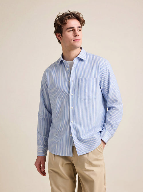 Man wearing a blue striped merino shirt with long sleeves and chest pocket on a beige pants against a beige background