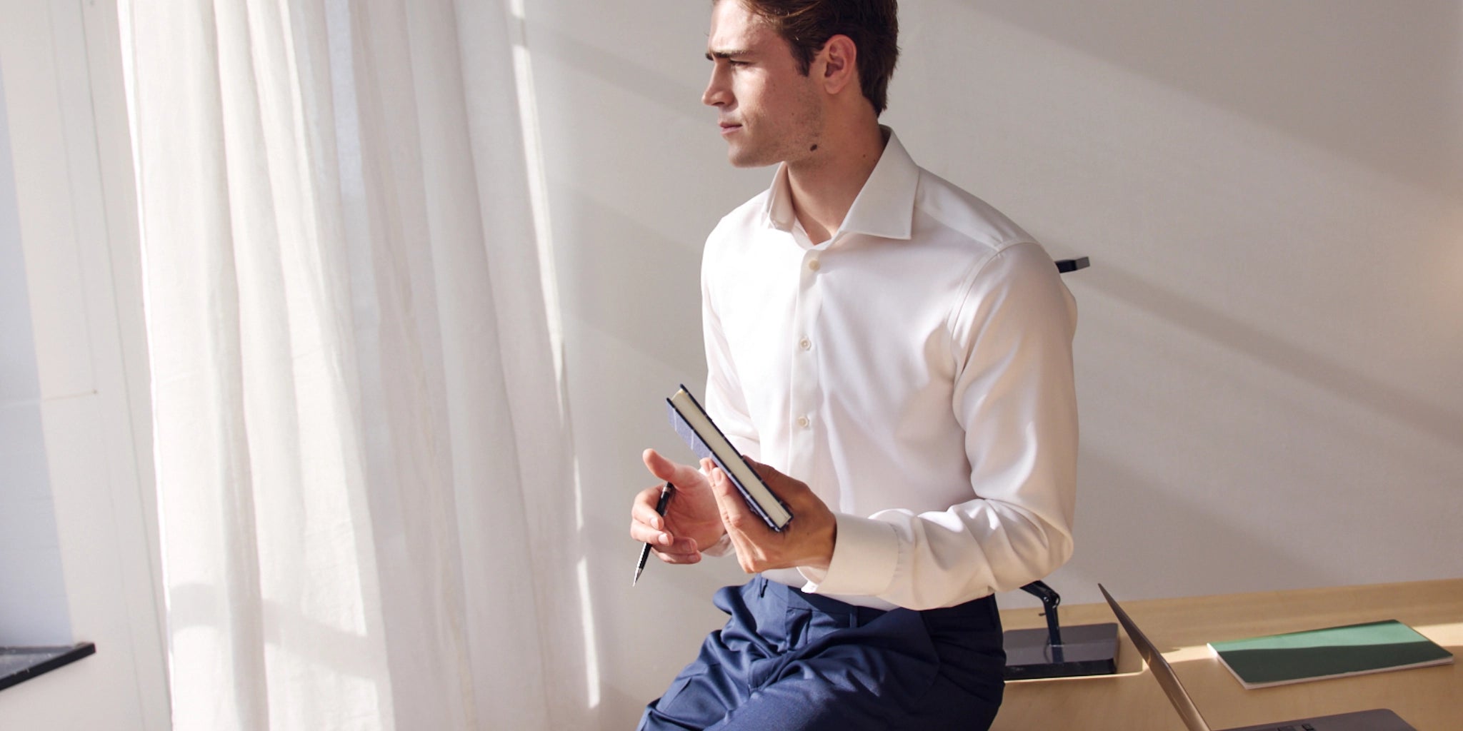 Man in a white merino dress shirt holding a notebook  in a bright office with white curtains.