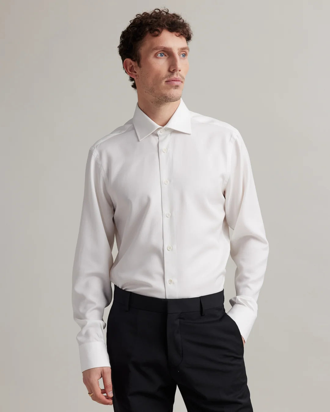 Man wearing a white dress shirt in merino wool with spread collar and black pants against a plain background