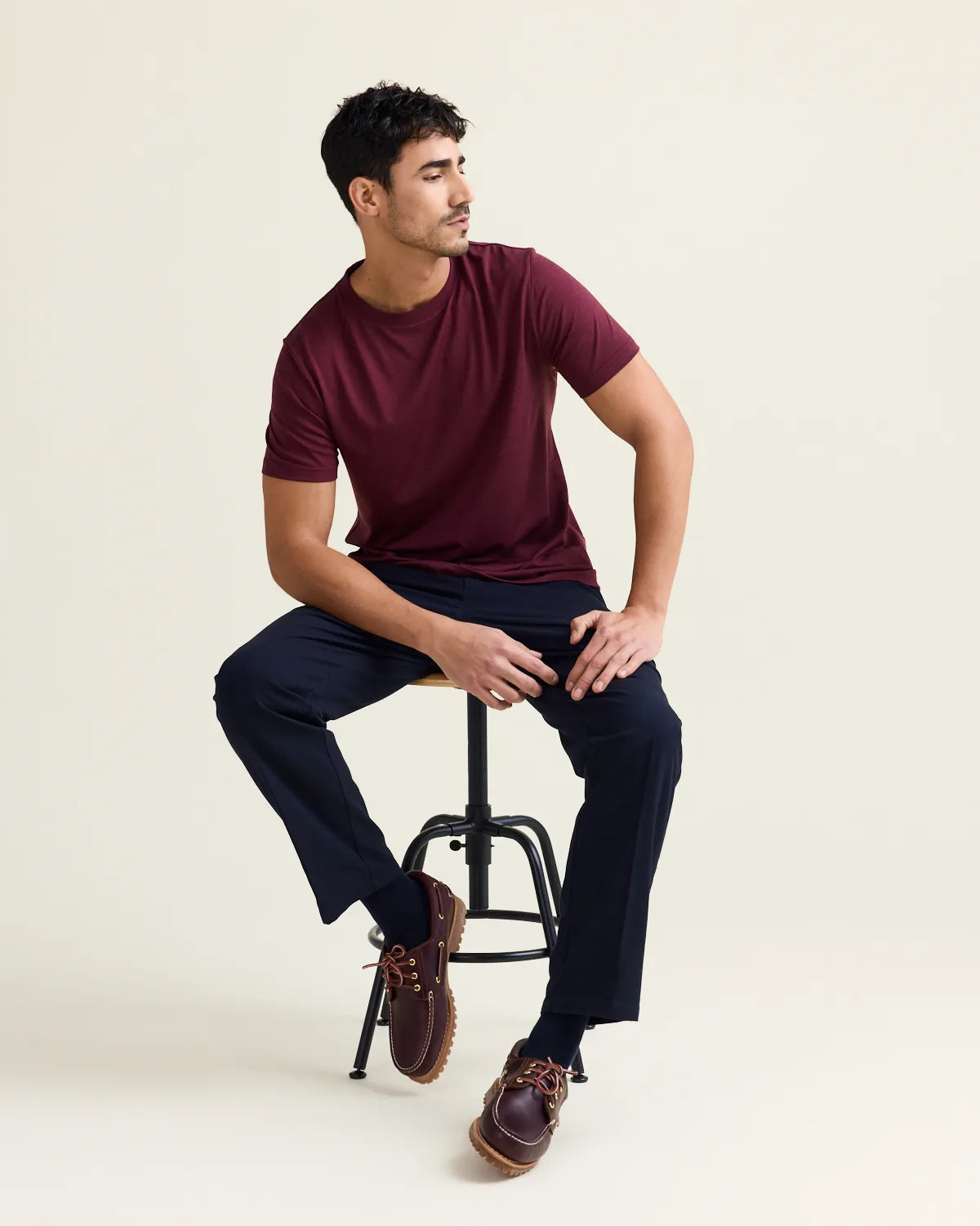 Man sitting on a stool wearing a burgundy  merinot-shirt and dark pants against a beige background