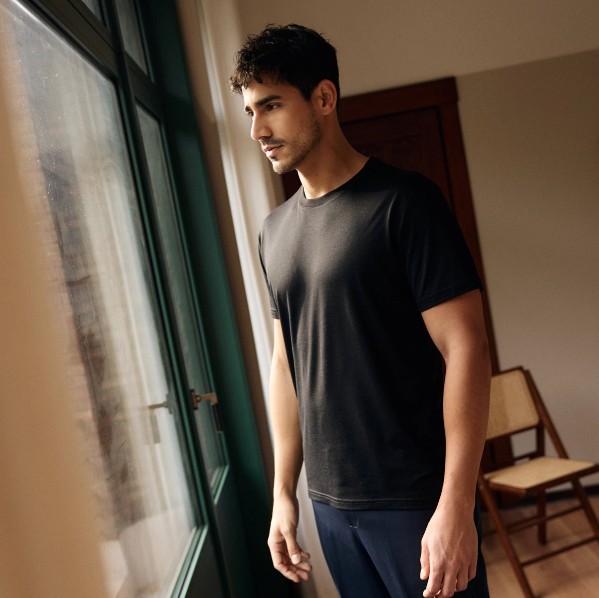 Man wearing a black merino wool T-shirt and is standing by a window in a room with a chair in the background