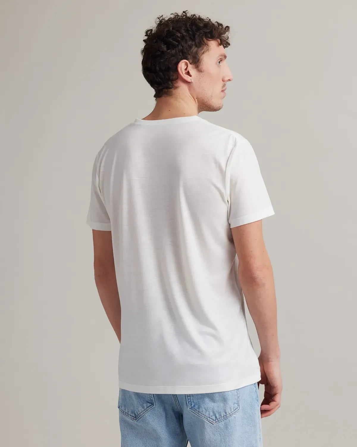 Back view of a man wearing a plain white merinowool t-shirt and light blue jeans against a neutral background