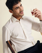 Man in a white merino dress shirt sits on a wooden chair with  a cocktail against a beige background
