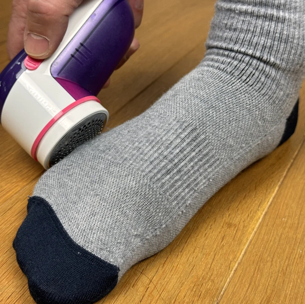 Person using a fabric shaver on a gray merino wool sock on a wooden floor.