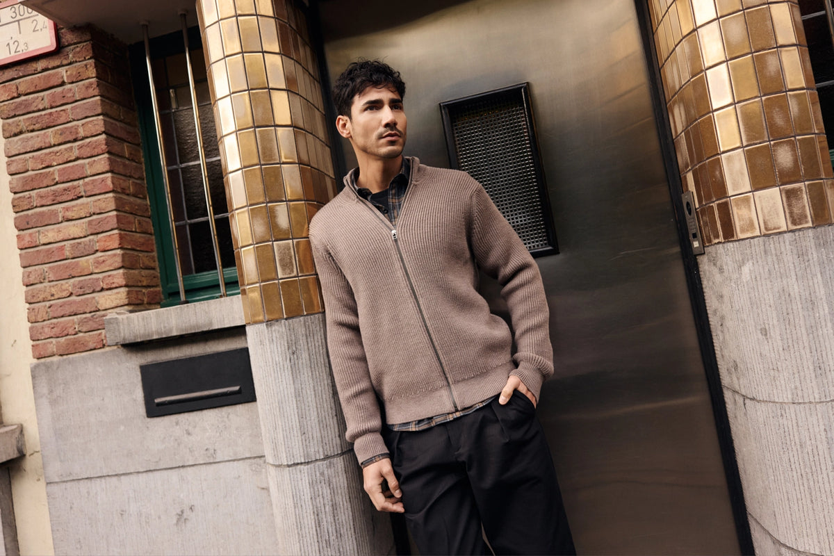 Man standing against a building with decorative columns in a wolk merino wool cardigan in brown colour with a flannel shirt in checks