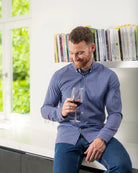 Man in a blue checkered merino long sleeve shirt holding a glass of red wine in a room with a window and bookshelf.
