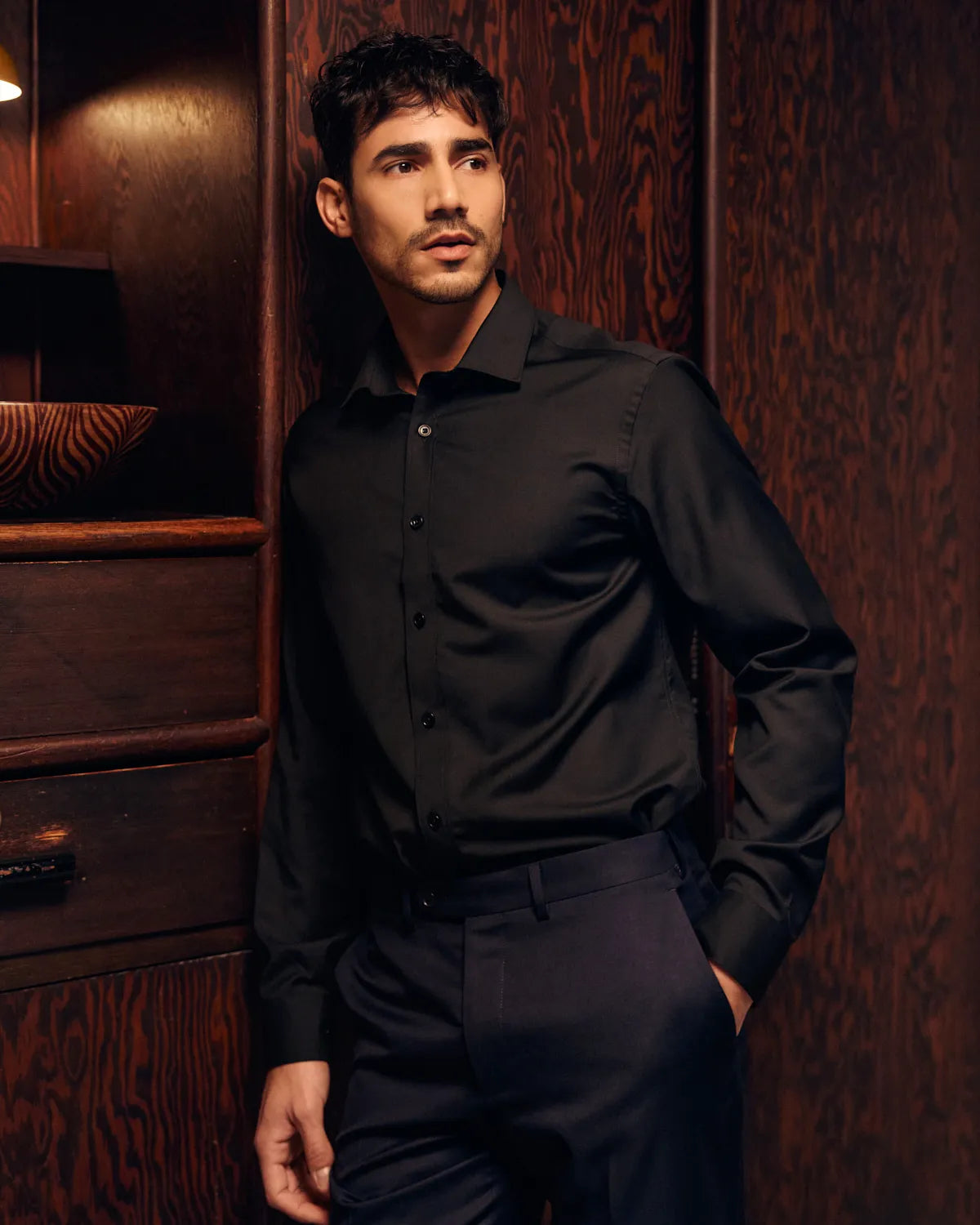 Man wearing a black merino dress shirt and pants against a wooden wall.