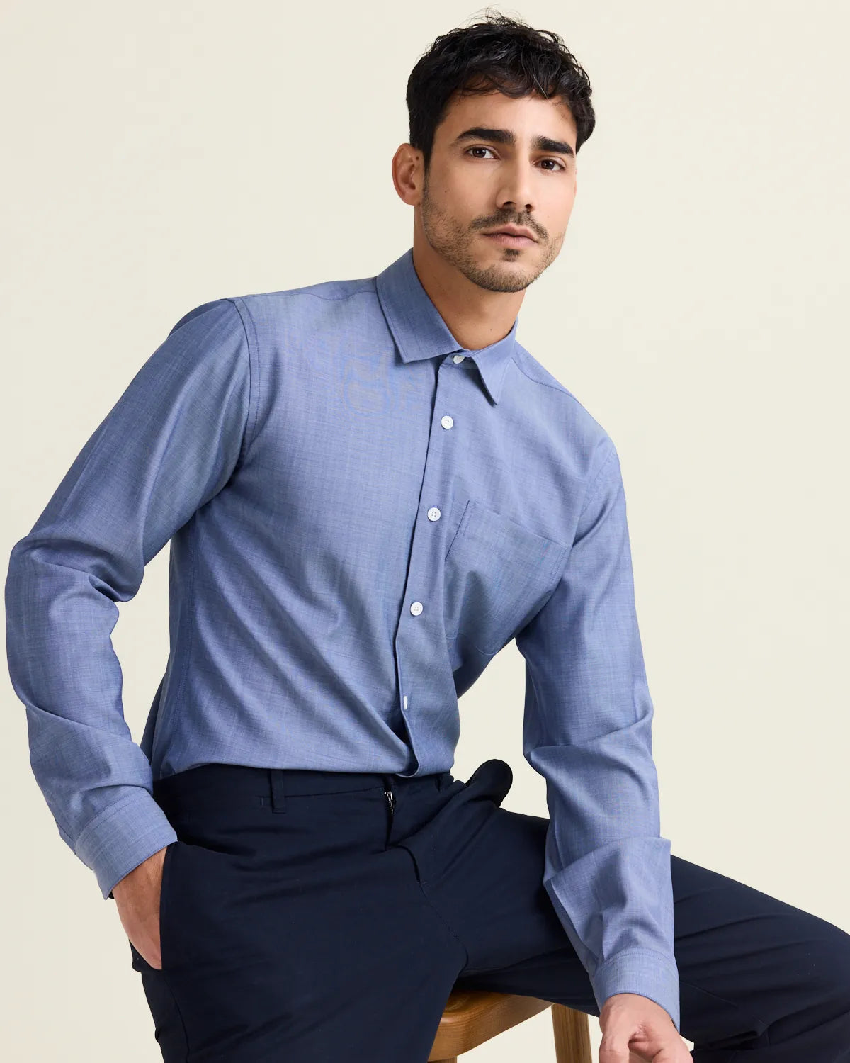 man on a stool wearing a light blue denim merinowool shirt and dark navy trousers on a plain background