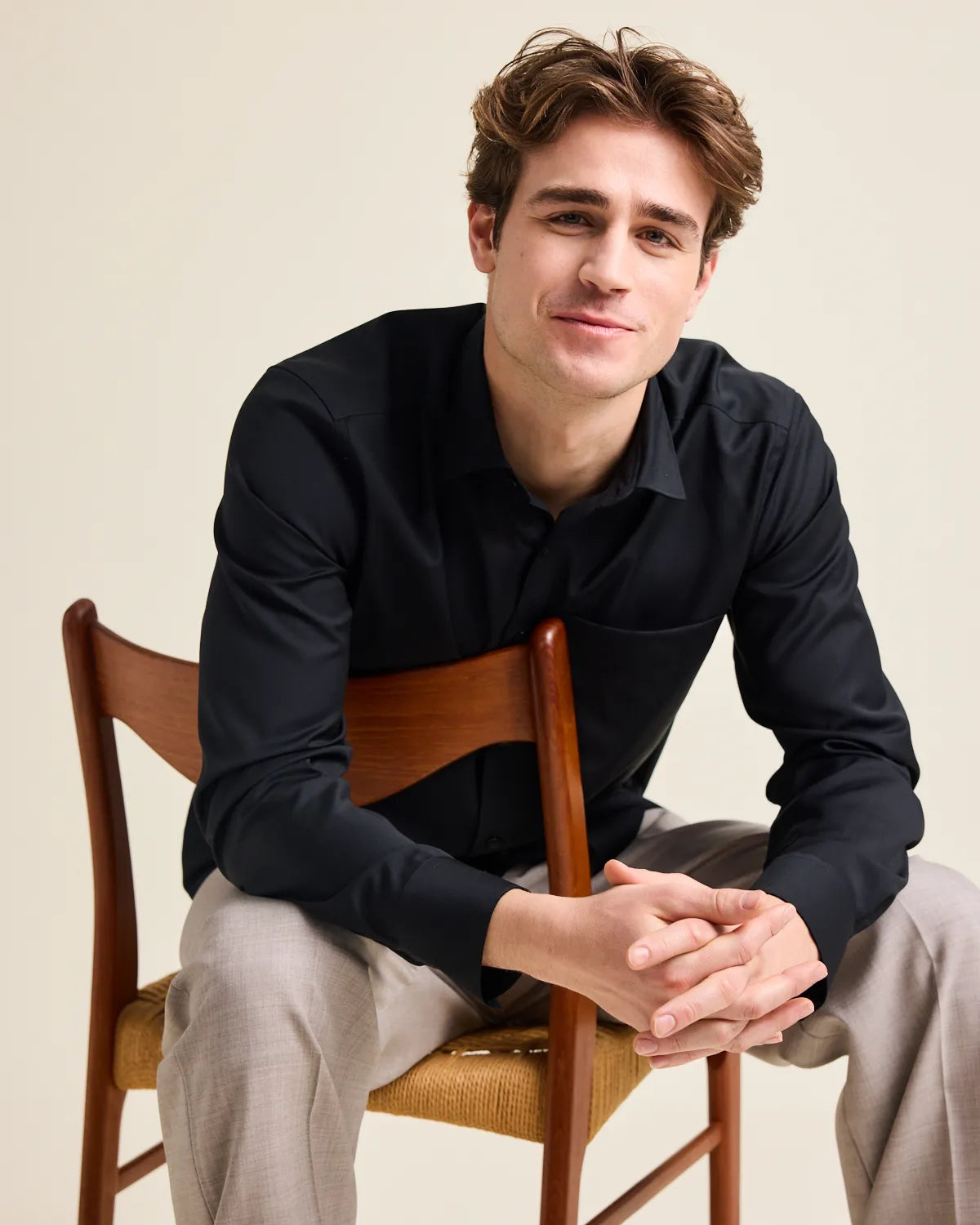 Man sitting on a wooden chair wearing a black merinowool shirt and light gray pants against a beige background