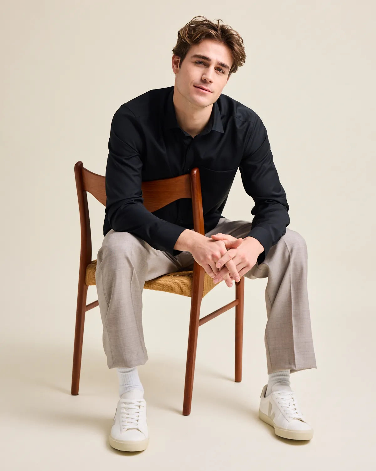 Man sitting on a wooden chair wearing a black merinowool button up shirt, gray pants, and white sneakers.