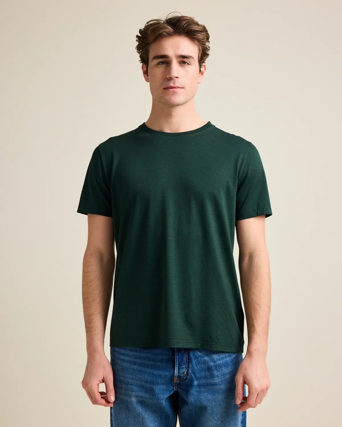 Man wearing a dark green t-shirt and blue jeans against a beige background