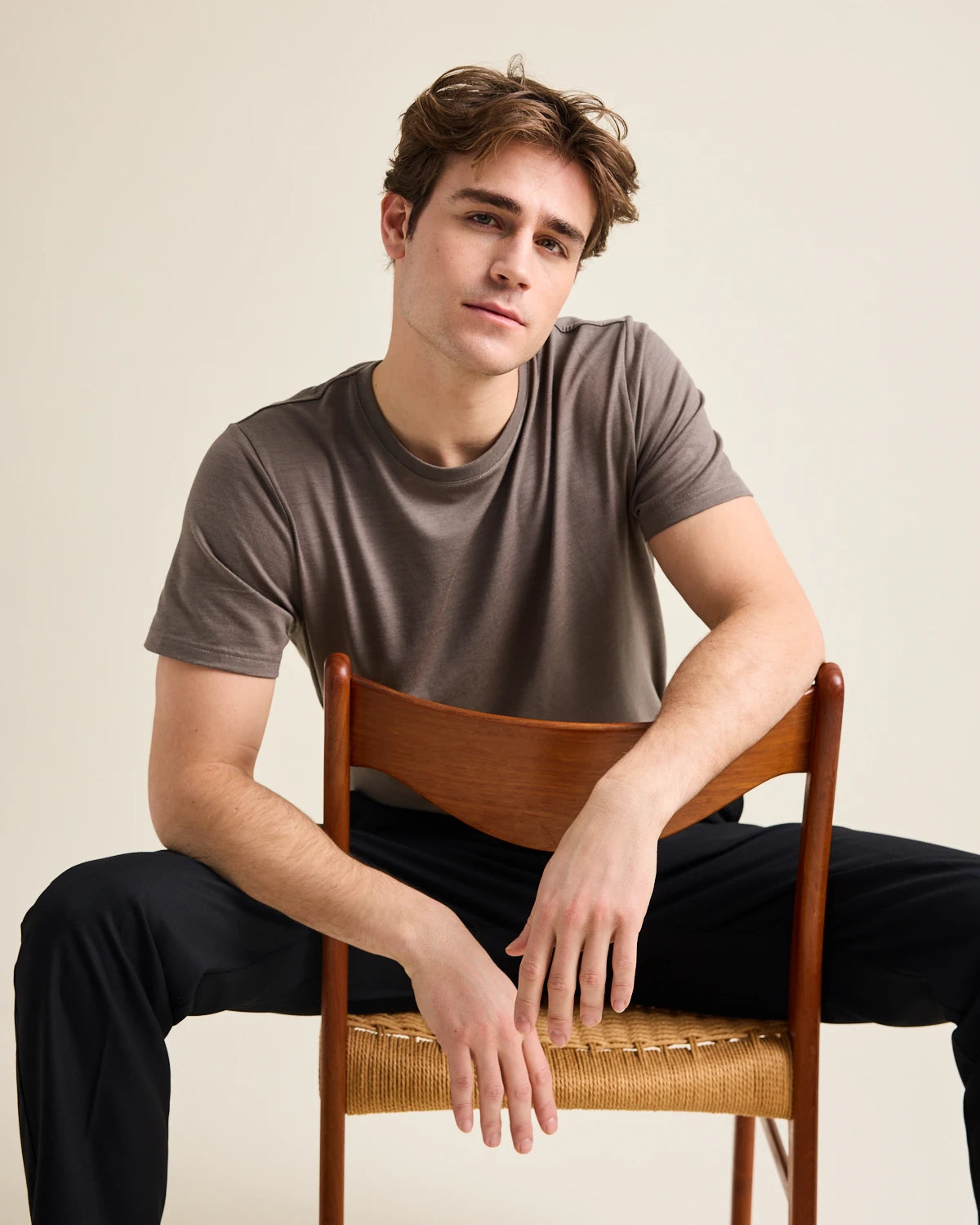 Man sitting on a wooden chair wearing a brown merino t-shirt and black pants against a beige background