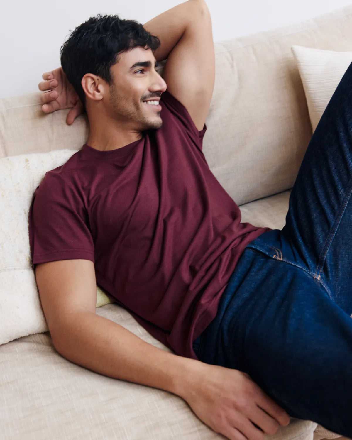 Man in a burgundy merino wool t-shirt from Wolk and blue jeans lying on a beige couch.