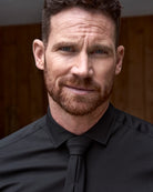 Man wearing a black merinowool shirt and a black tie against a wooden background