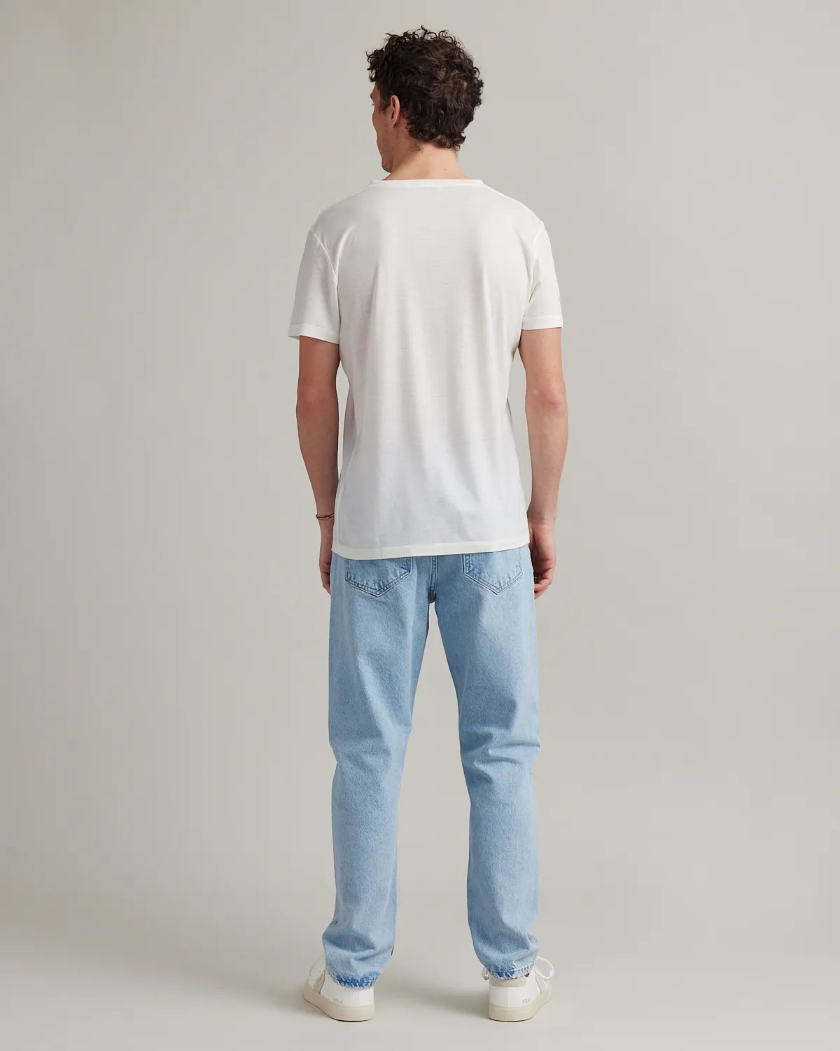 back view of a man wearing a white merino t-shirt and light blue jeans on a plain background