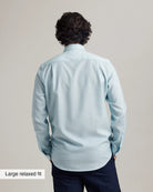 back view of a man with an untucked light blue merinowool dress shirt on dark navy trousers and a plain background