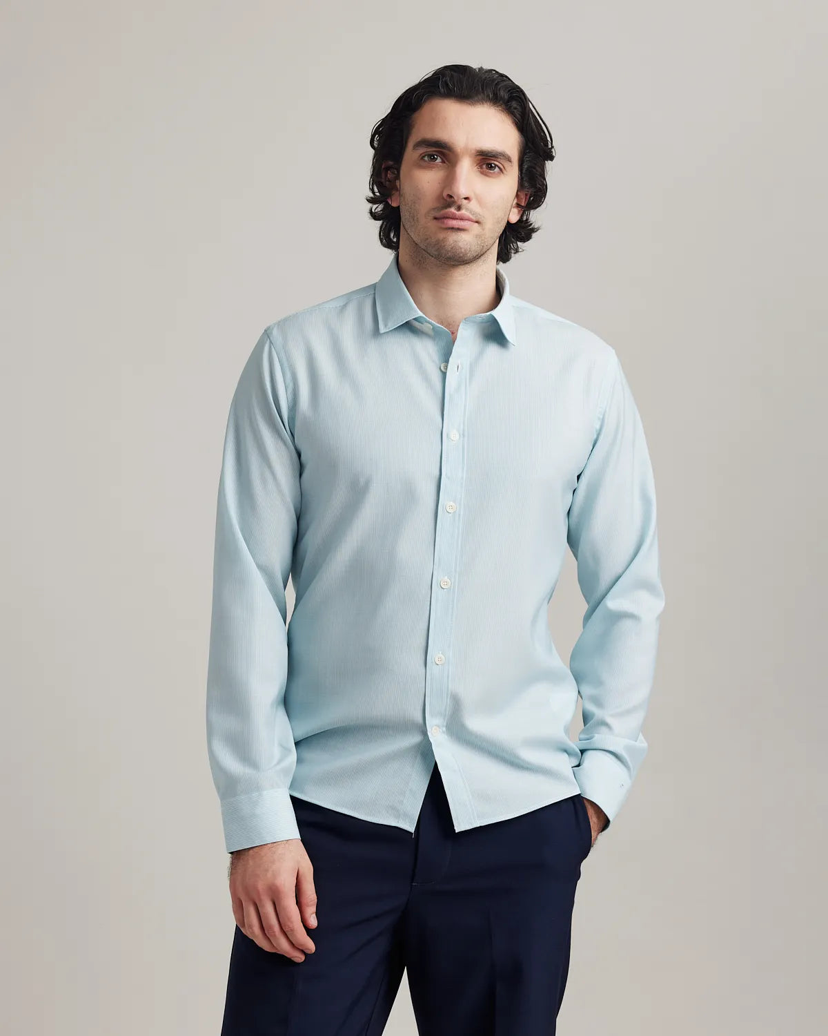 Man wearing a light blue merinowool dress shirt and dark pants against a plain background