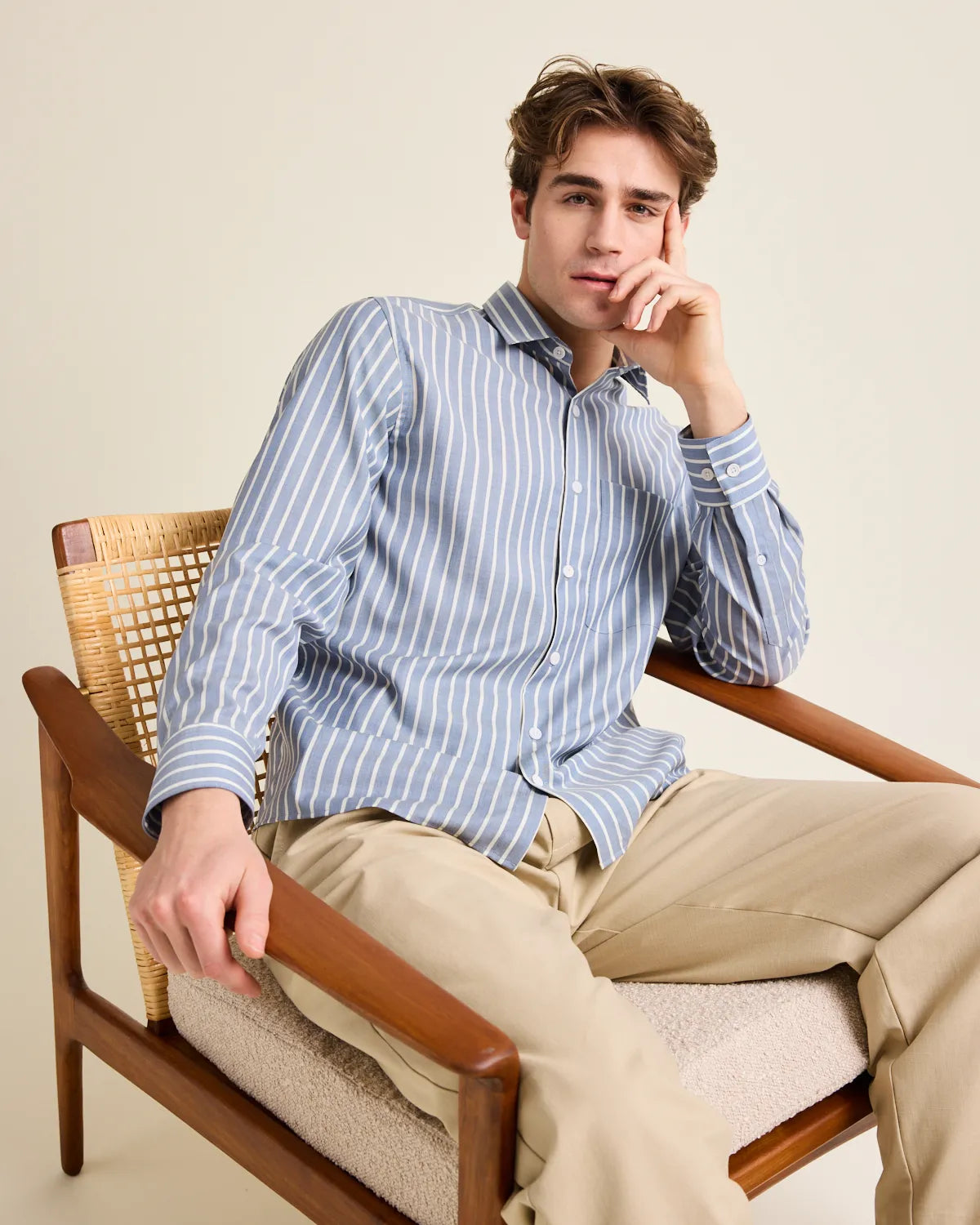 Man sitting in a wooden chair wearing a navy blue and white striped merino wool button up shirt  and beige pants on a plain background