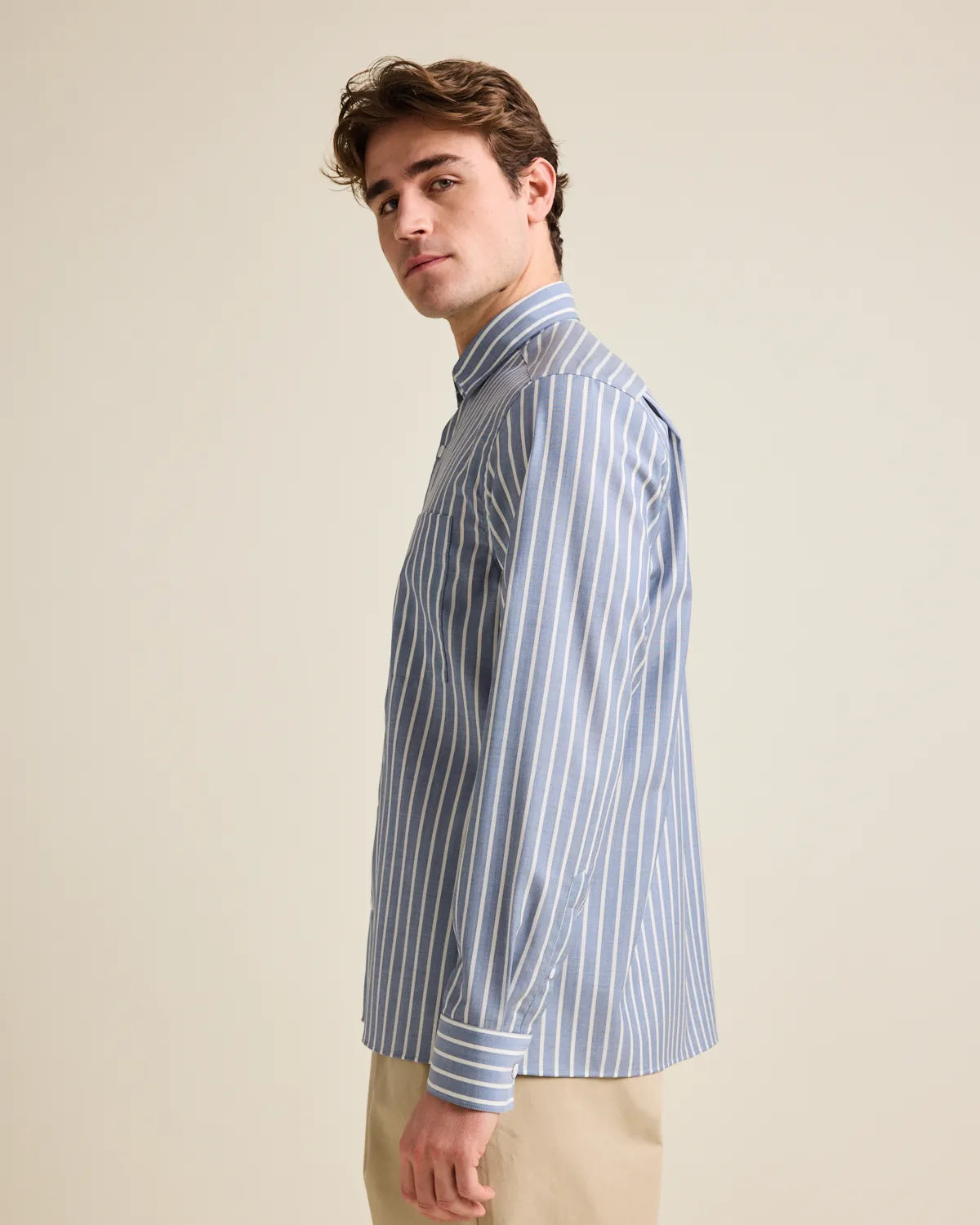Side view of a man wearing a navy blue and white striped merino wool button up shirt  against a beige background