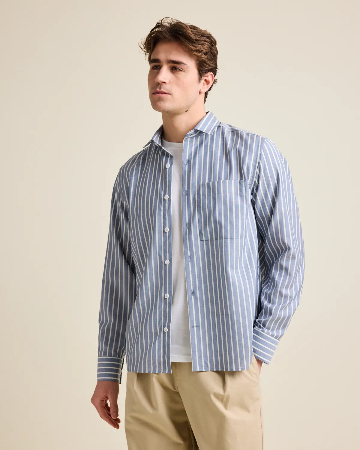 Man wearing a navy blue and white striped merino wool button up shirt over a white Tshirt with beige pants on a beige background