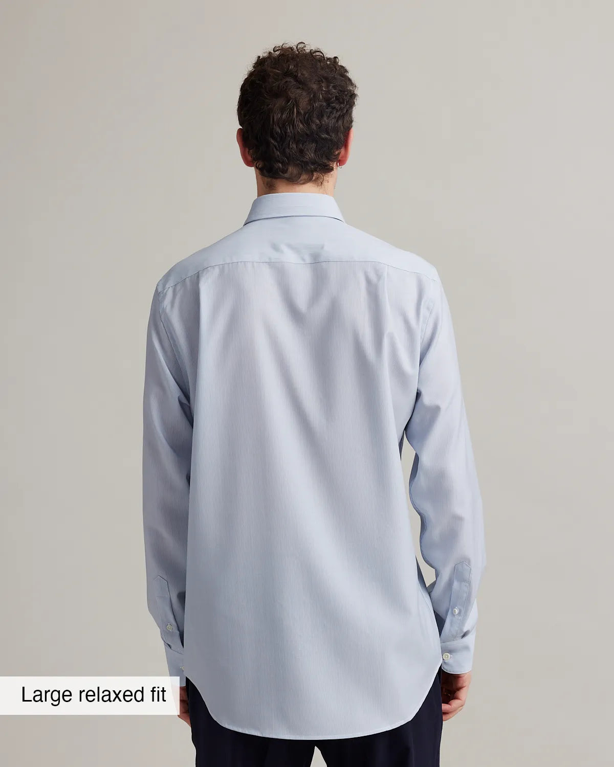 Back view of a man wearing a light blue striped merino shirt with a large relaxed fit label at the bottom.
