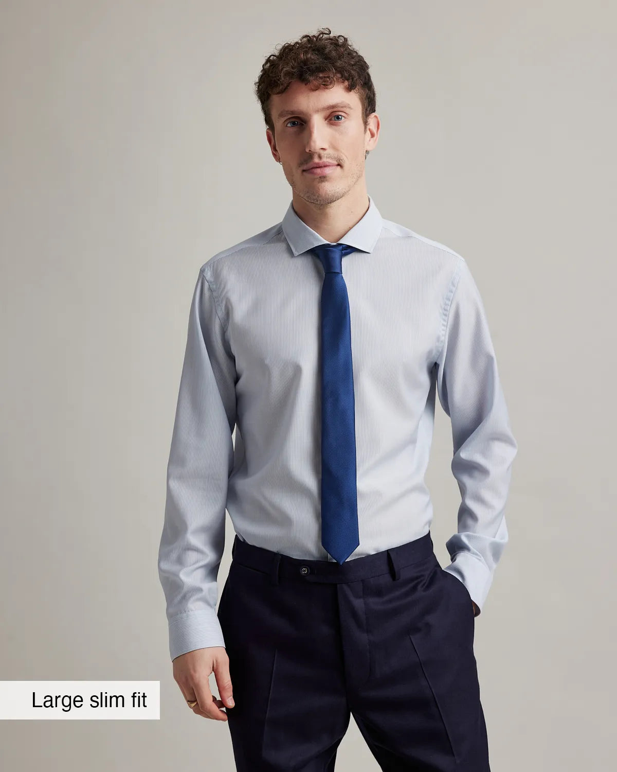 Man wearing a light gray merino wool dress shirt with a blue tie and dark pants on a plain background