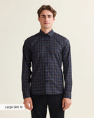 A man modeling a long-sleeve plaid Wolk merino shirt in a dark navy and black colorway, paired with black trousers, against a light beige background. A text overlay in the bottom-left corner reads 'Large slim fit'.