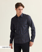 A man wearing a dark navy and blue plaid long-sleeve button-up merino wool shirt with black trousers, standing against a light beige background. A text overlay in the bottom-left corner reads 'Large slim fit'.