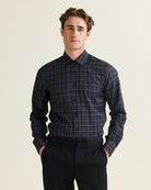 A man wearing a long-sleeve button-up plaid merino shirt in navy, purple and blue with thin grey and black lines, paired with dark trousers, standing against a light beige background.