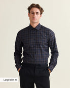A man wearing a dark navy and black plaid long-sleeve dress Wolk merino shirt with a slim fit, paired with black trousers, against a light beige background. A text overlay in the bottom-left corner reads 'Large slim fit'.