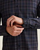 Close-up of a man's hands buttoning the cuffs of a long-sleeve plaid merino shirt in navy, blue, and gray.