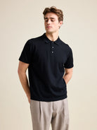Man wears a black merino polo shirt with short sleeves in a studio setting. 