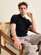 Man sits in vintage chair with black merino wool polo kintted from Cashwool Zegna yarn.