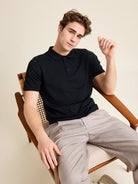Male model sits in vintage chair wearing a black merino wool polo shirt highlighting the quality of the Cashwool Zegna yarn.