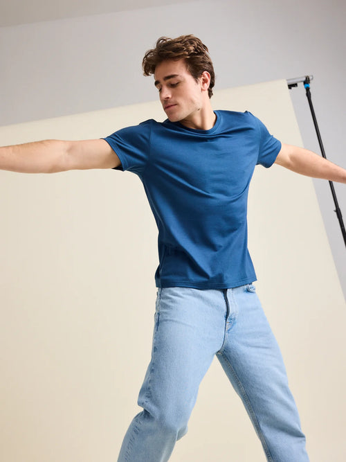 Studio shot of a men's denim blue colored merino wool T-shirt from Wolk, worn by a model that jumps in front of the camera. 