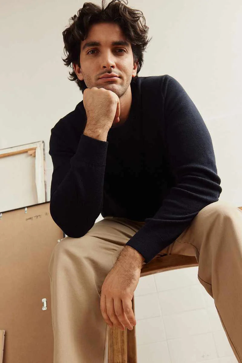 Man sitting on a chair wearing a dark navy merino wool sweater and beige pants against a neutral background