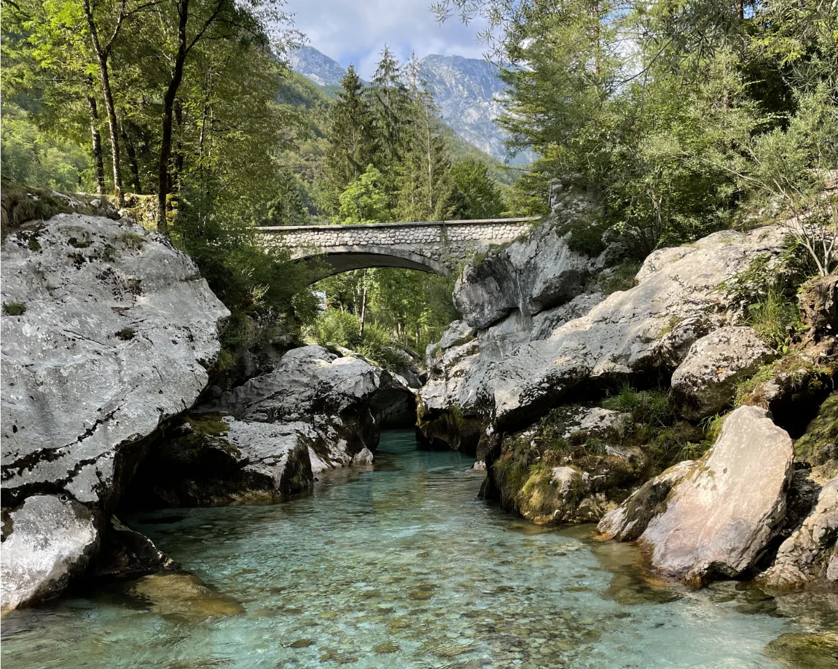 Stone bridge over a clear stream with rocky cliffs and trees in the background